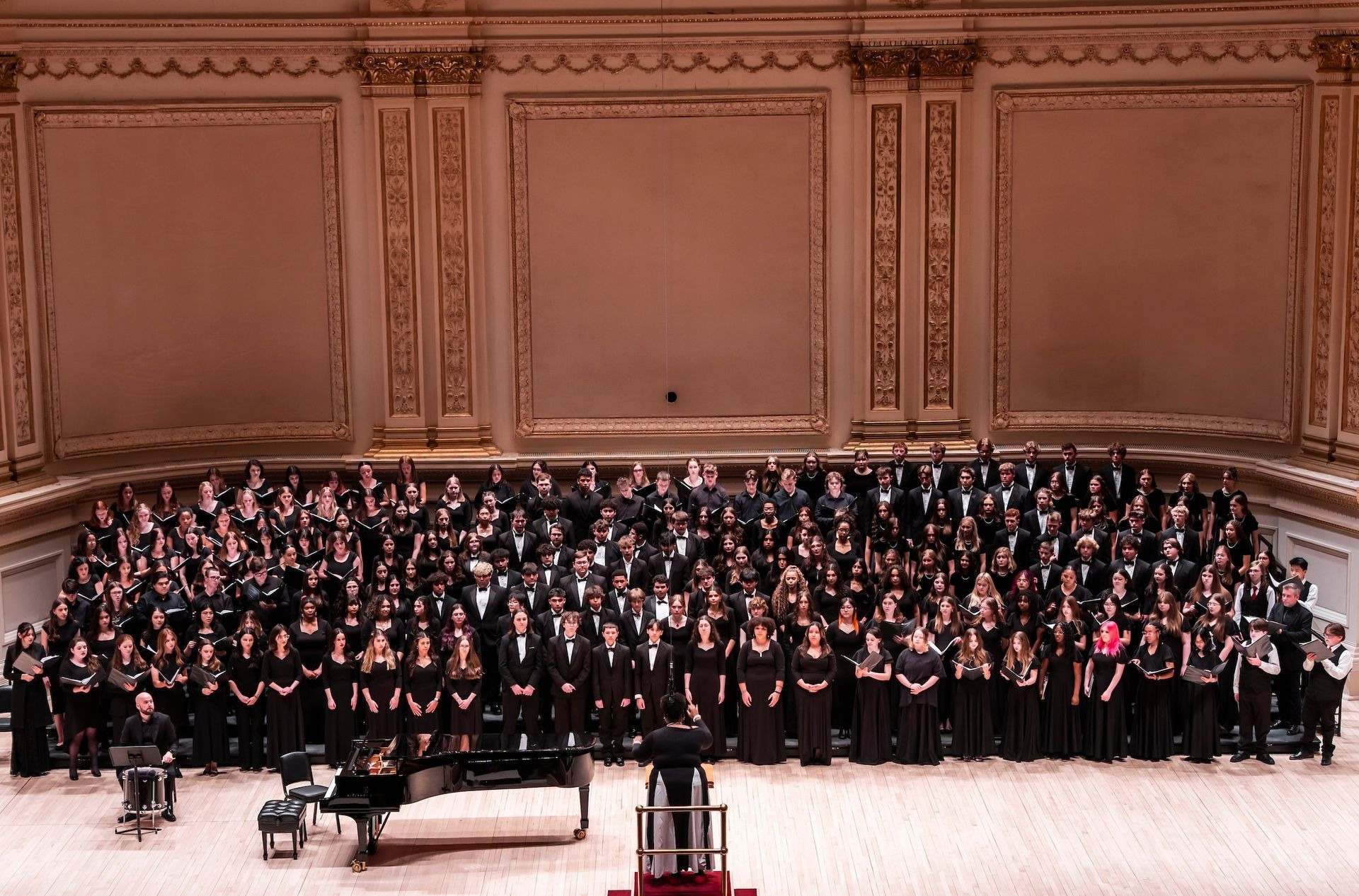 A massed choir in black formal dress stands on the Carnegie Hall stage, a conductor at the podium and a grand piano to the left.