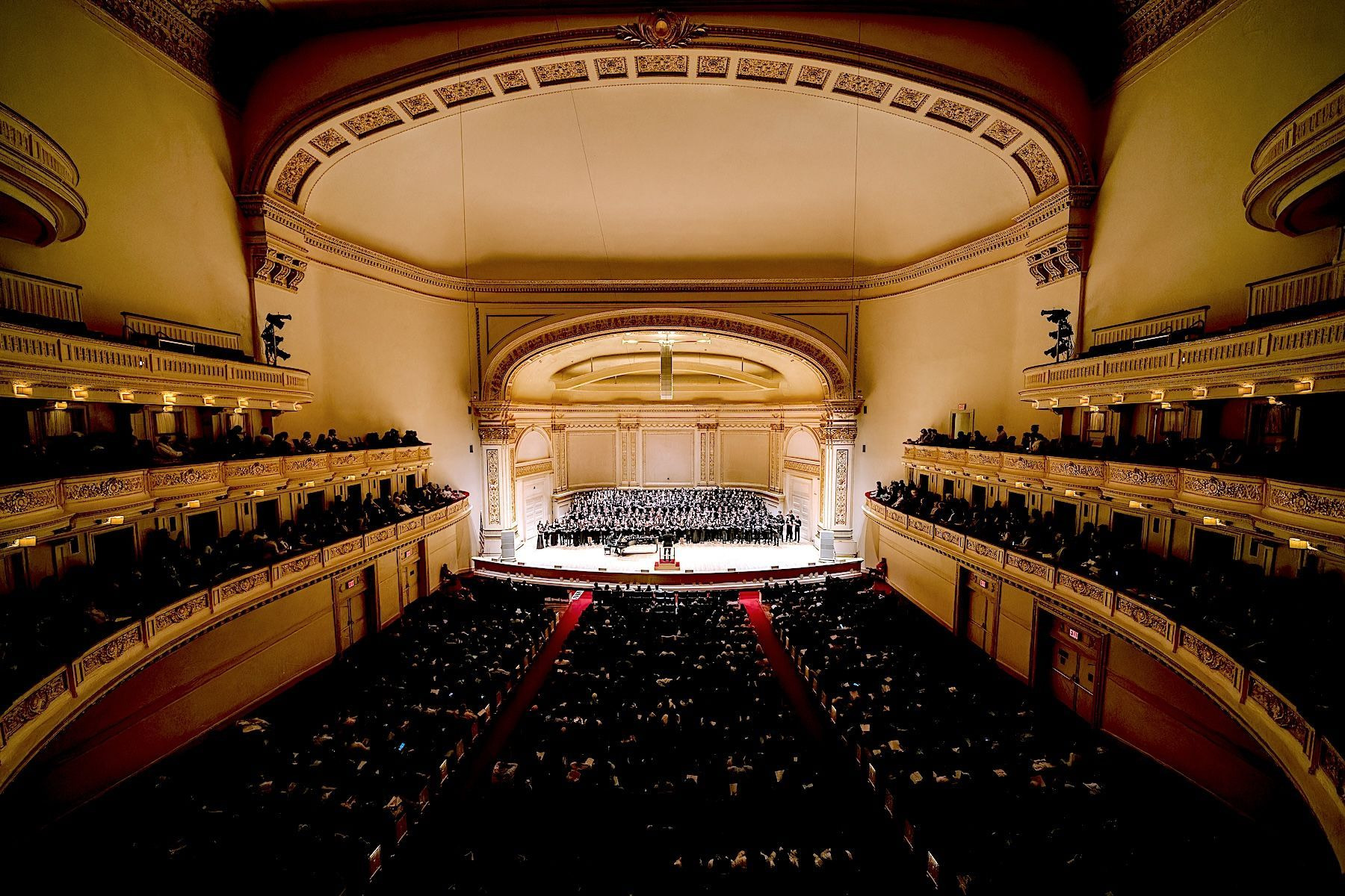 From a high balcony at Carnegie Hall, a choir in black is lit on the stage while the house and side balconies sit in the dark.