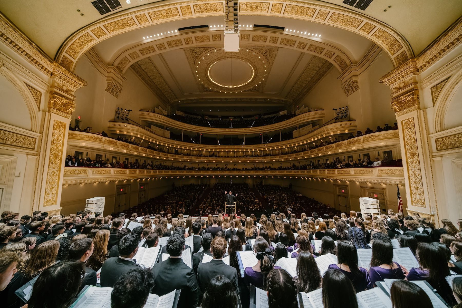 From within a massed choir at Carnegie Hall, singers hold music and face a conductor, with a full house and gilded balconies beyond.