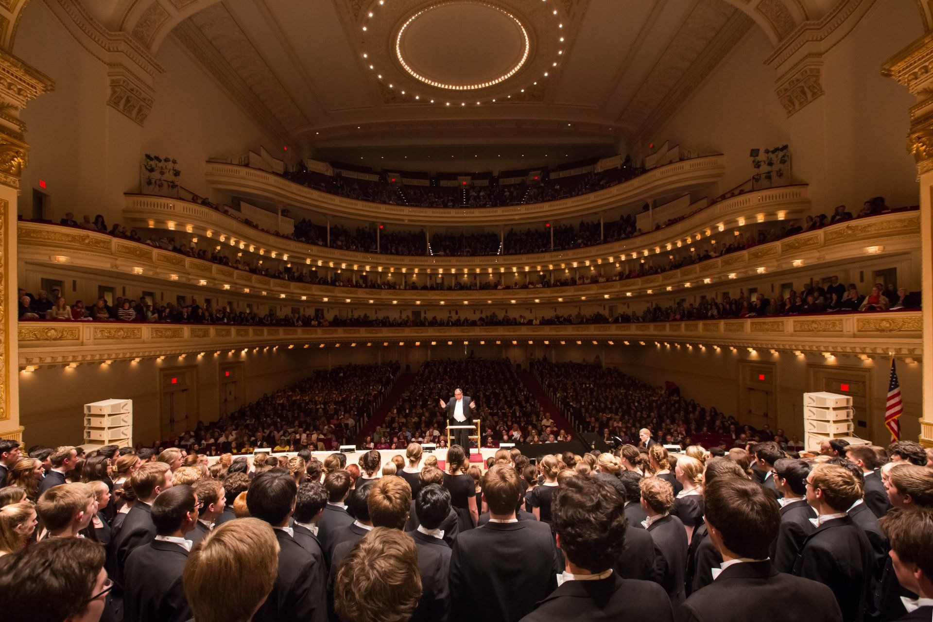 Choirs of America at Carnegie Hall, seen from the stage: singers face a packed house under a ring of ceiling lights.
