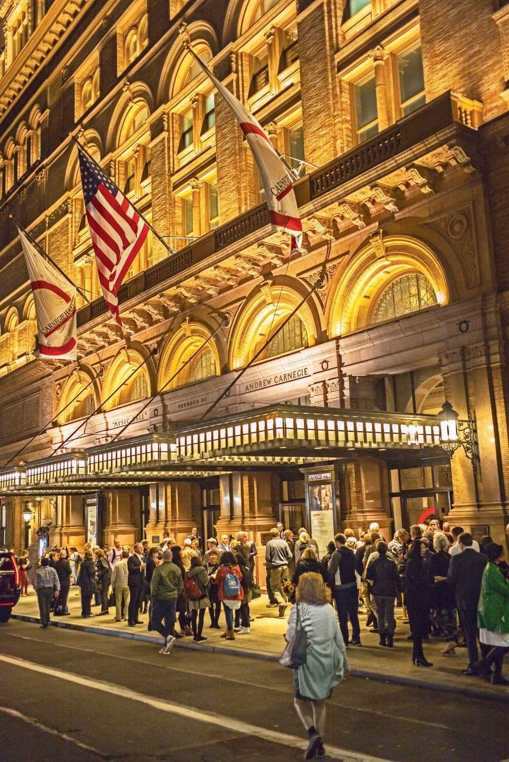 Carnegie Hall’s entrance at night, marquee lit, flags above a crowd on the sidewalk.