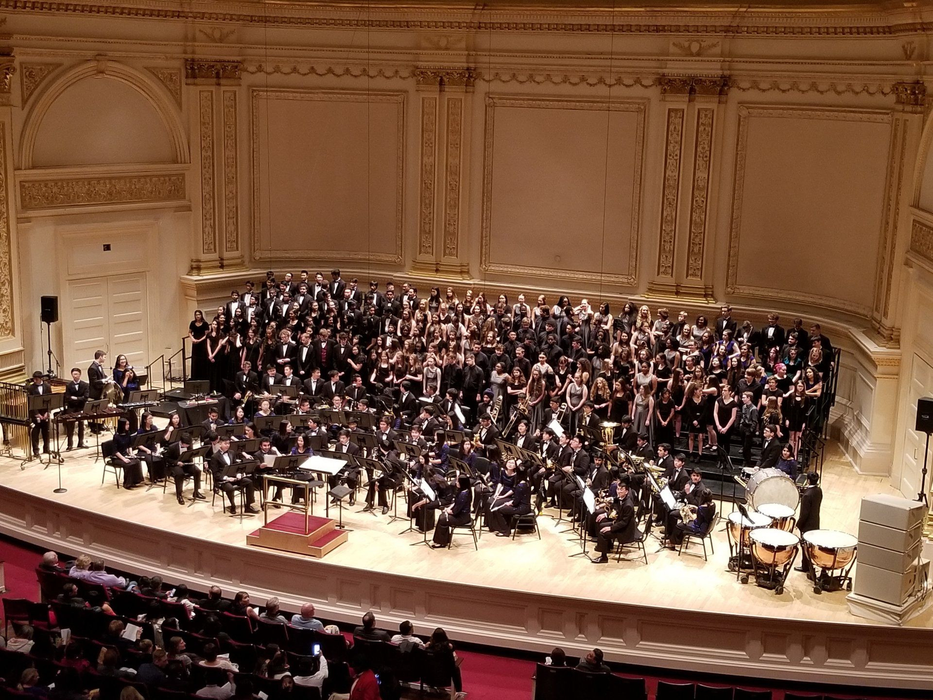 An orchestra and a massed choir share the Carnegie Hall stage, timpani at the right, audience in the foreground.