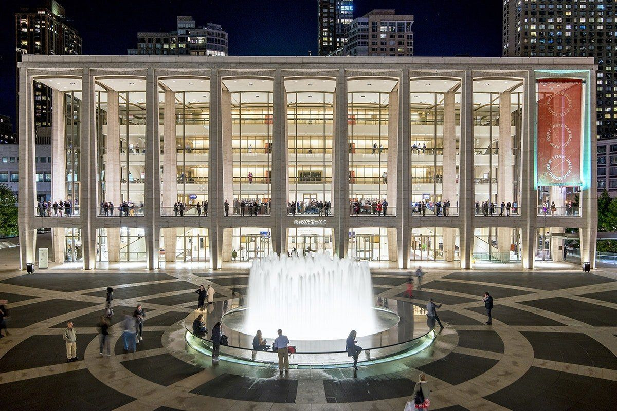 David Geffen Hall at Lincoln Center at night, the plaza fountain lit, people on the lobby levels.