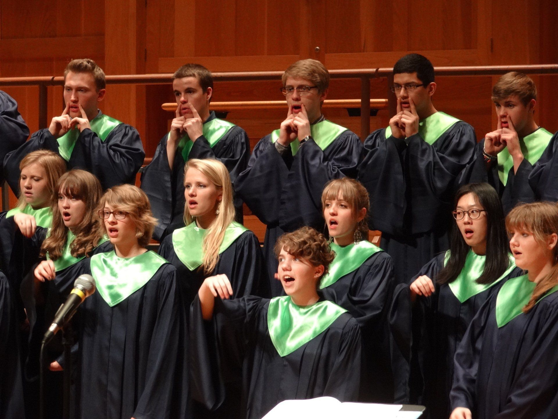 Young singers in navy robes and green stoles rehearse in a wood-paneled room.