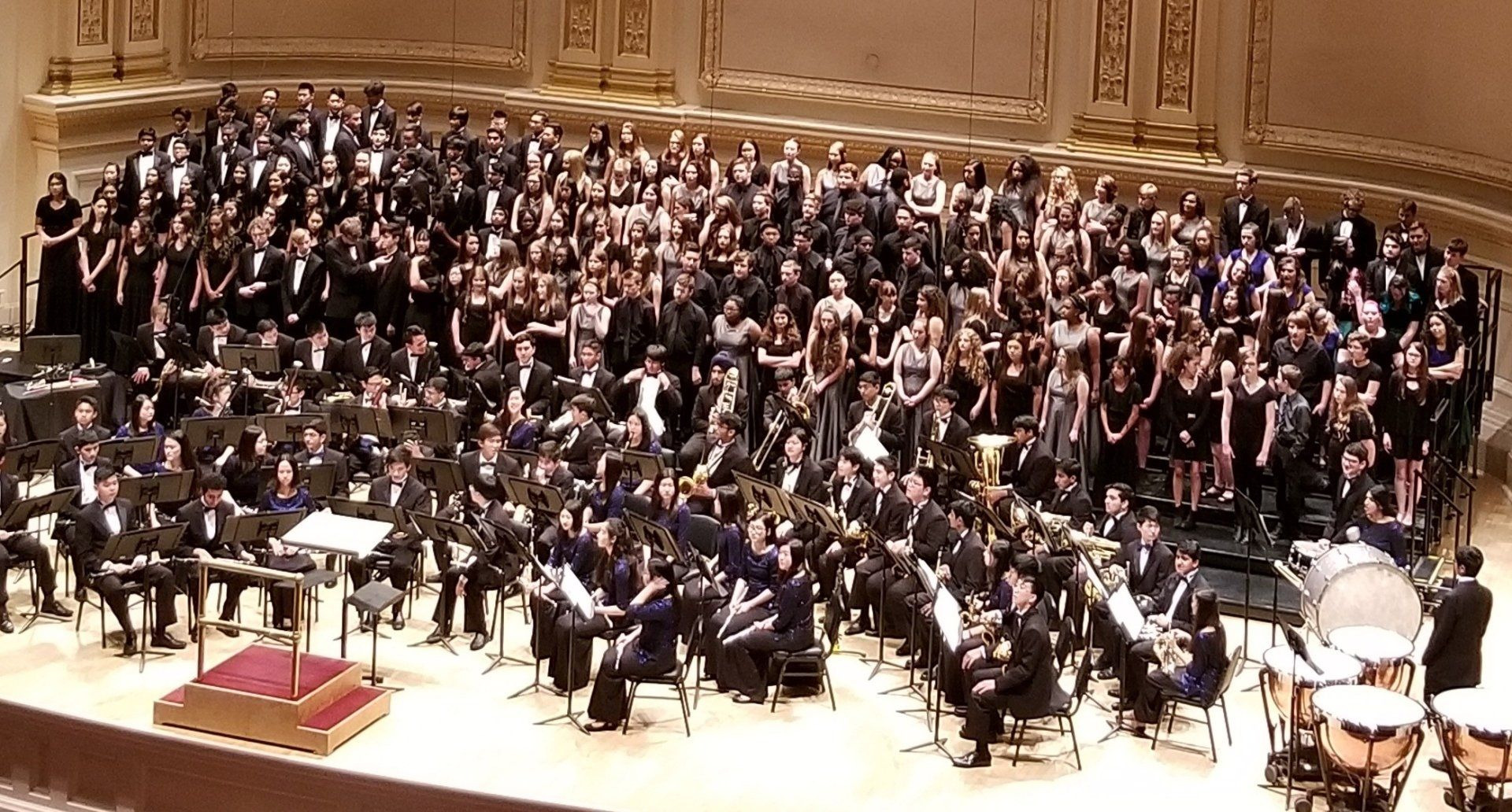 A band and a massed choir share a concert stage, timpani in the foreground.