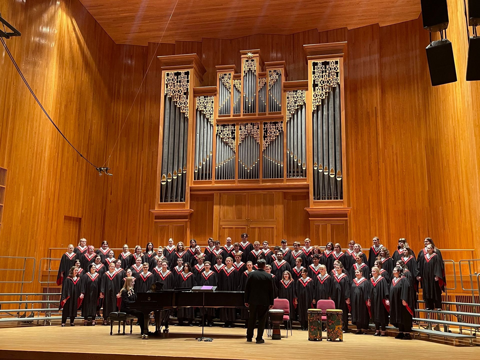 A choir in black robes with red stoles stands before a pipe organ while a conductor faces them.